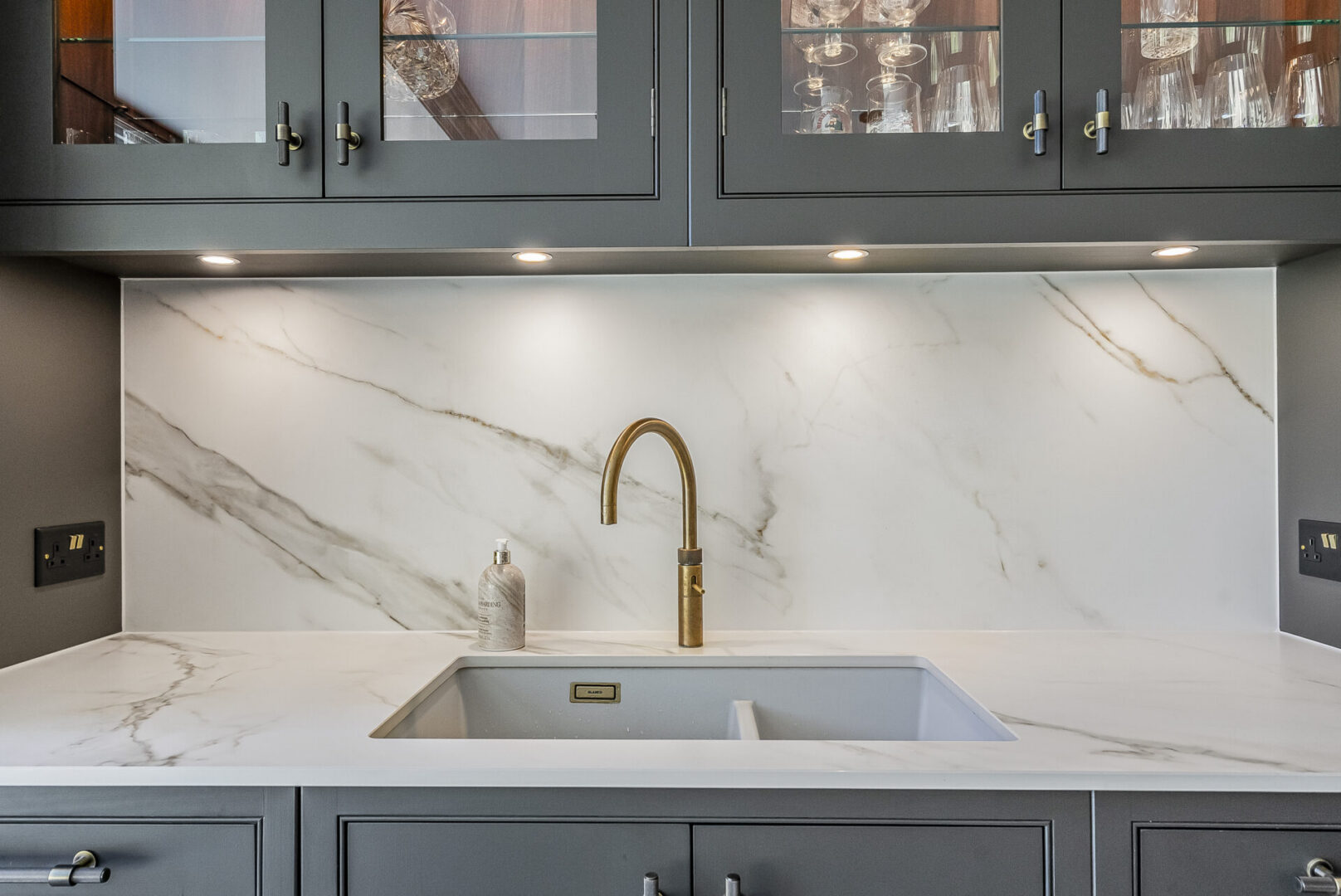 Kitchen with a white marble countertop, a sink, a tap, a bottle, cupboards, lights, and glasses in the cupboard