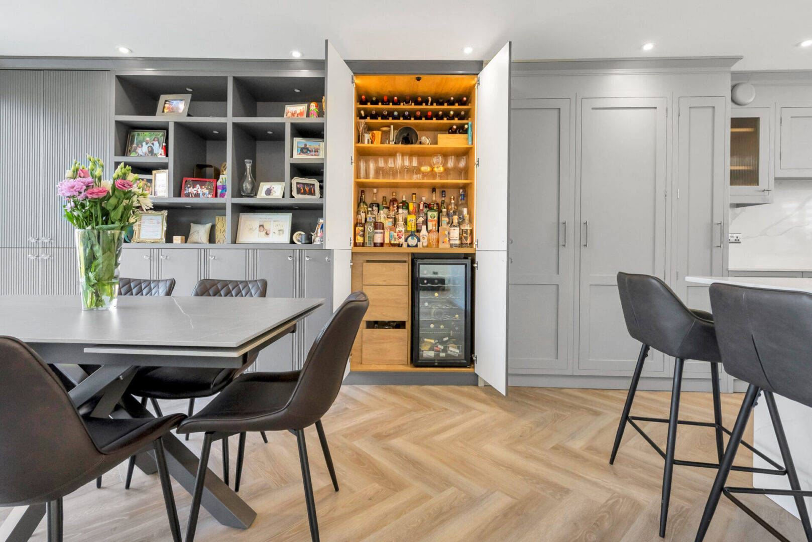 Dining room with a table and chairs, a flower vase on the table, cupboards, racks with bottles, photo frames, vases