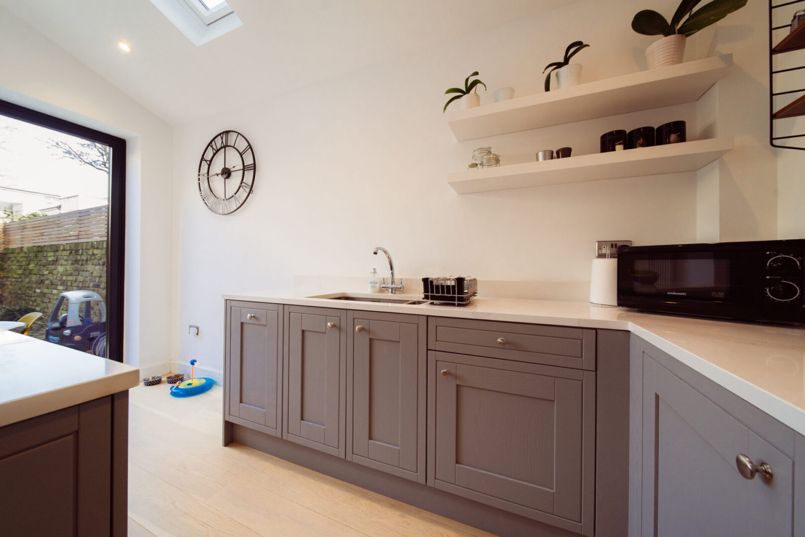 Kitchen with a sink, tap, microwave oven, cupboards, clock on the wall, houseplants, crockery, electric lights to the roof
