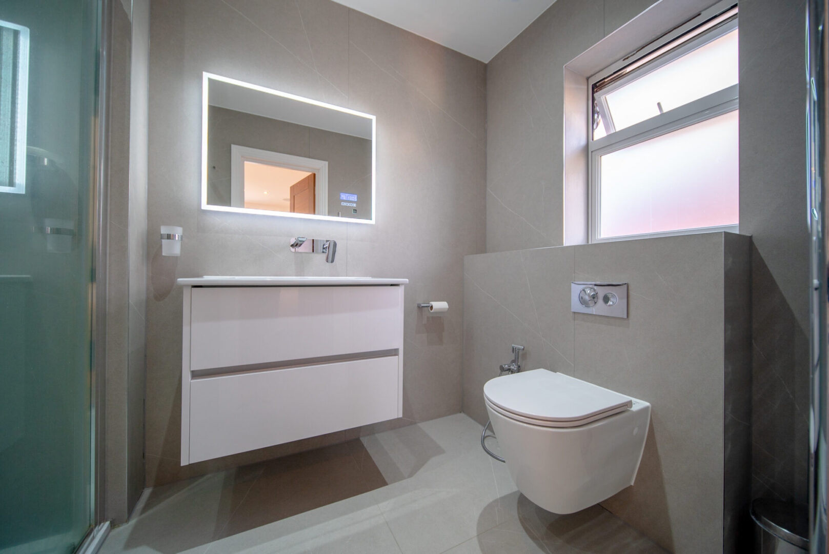 Bathroom with a toilet, sink, mirror, flush tank, tissue paper roll, taps, cupboards, glass door, dustbin and a window